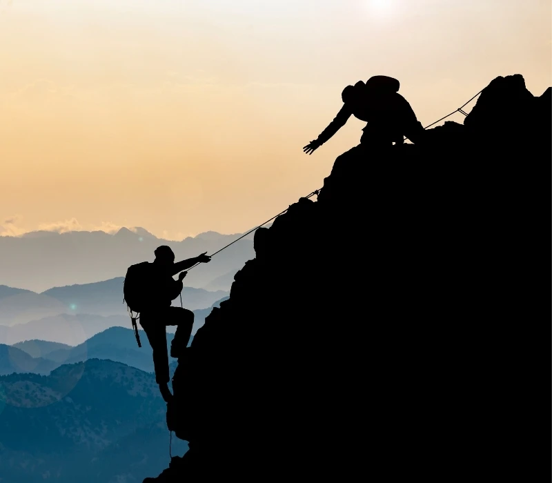 Two silhouettes of people climbing a mountain at sunset.